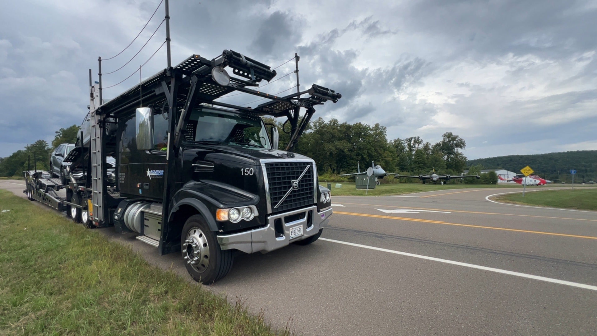 Black Allstar Hauling Volvo VHD car hauler loaded with SUVs parked beside vintage aircraft at an air park
