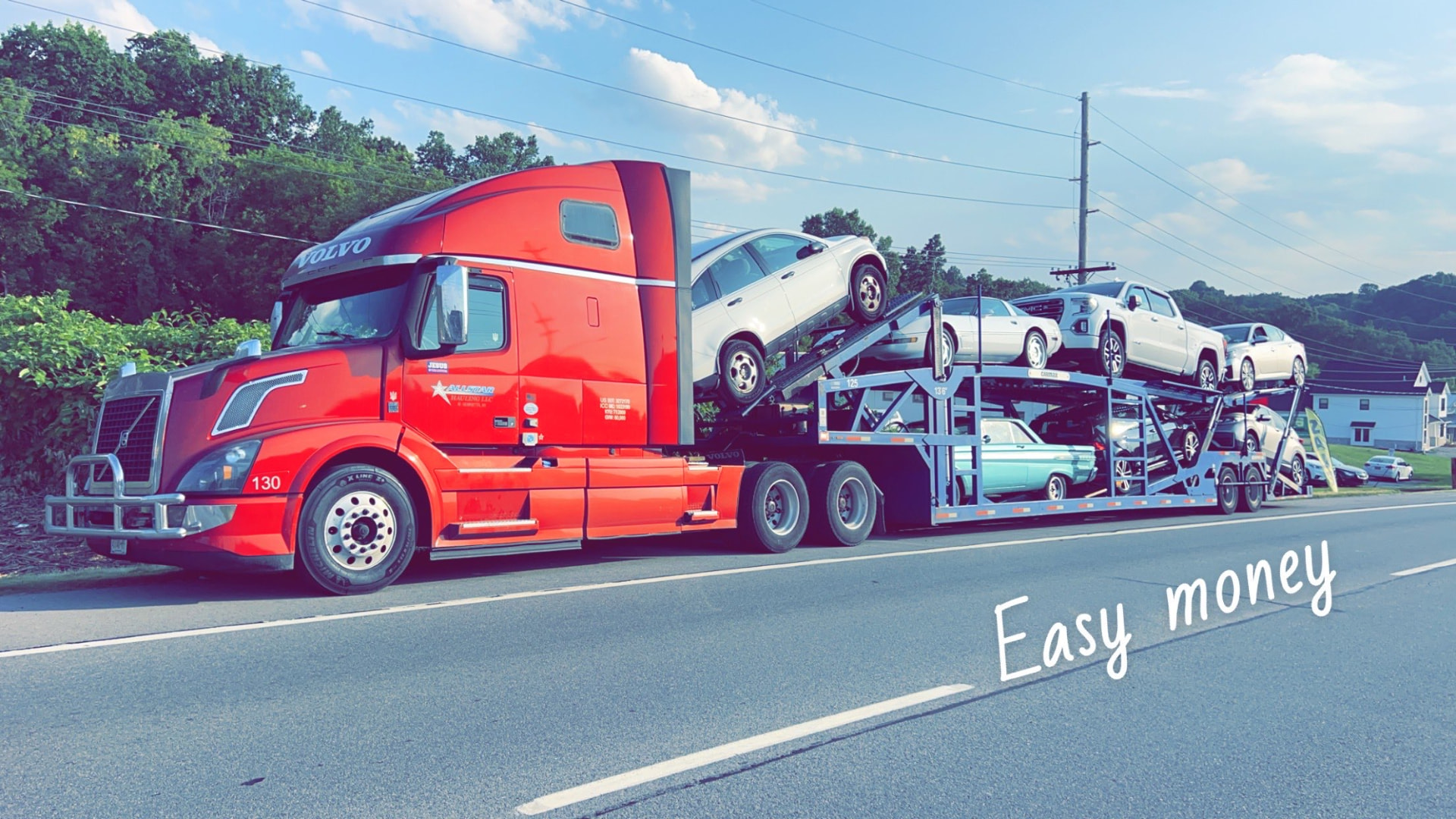 Red Allstar Hauling Volvo VNL car carrier loaded with SUVs and a classic Mercury rolling down a summer highway